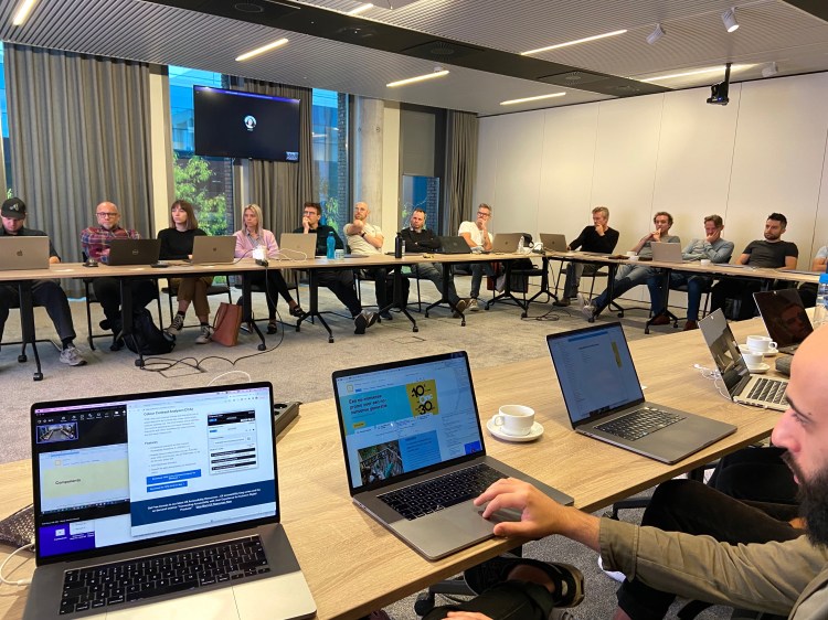 Group of 13 (visible) people attending the training with their laptops open, seating in a U table setup.