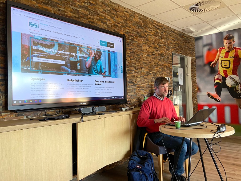 Trainer demoing screen reader on BASE website. Seating at a table with TV screen in the back.