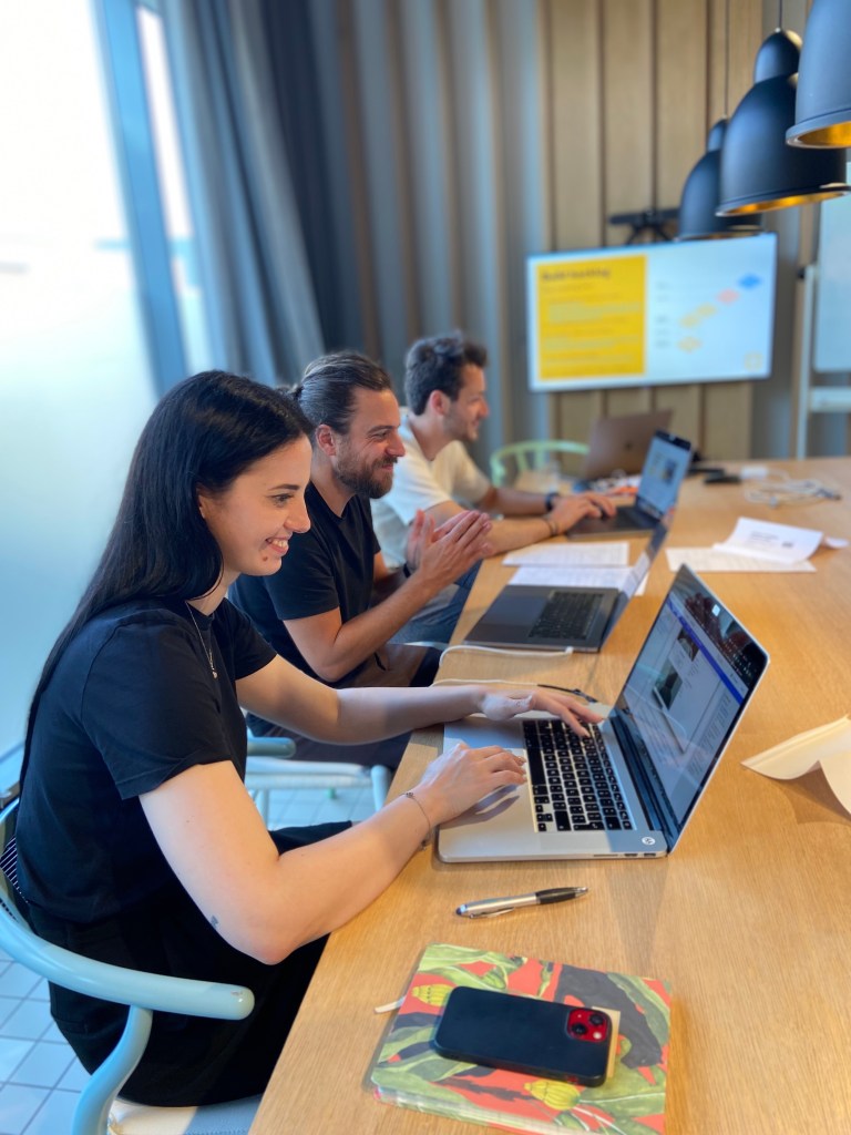 Tree people smiling seating in a row  at a table facing their laptops. Girl in foreground.