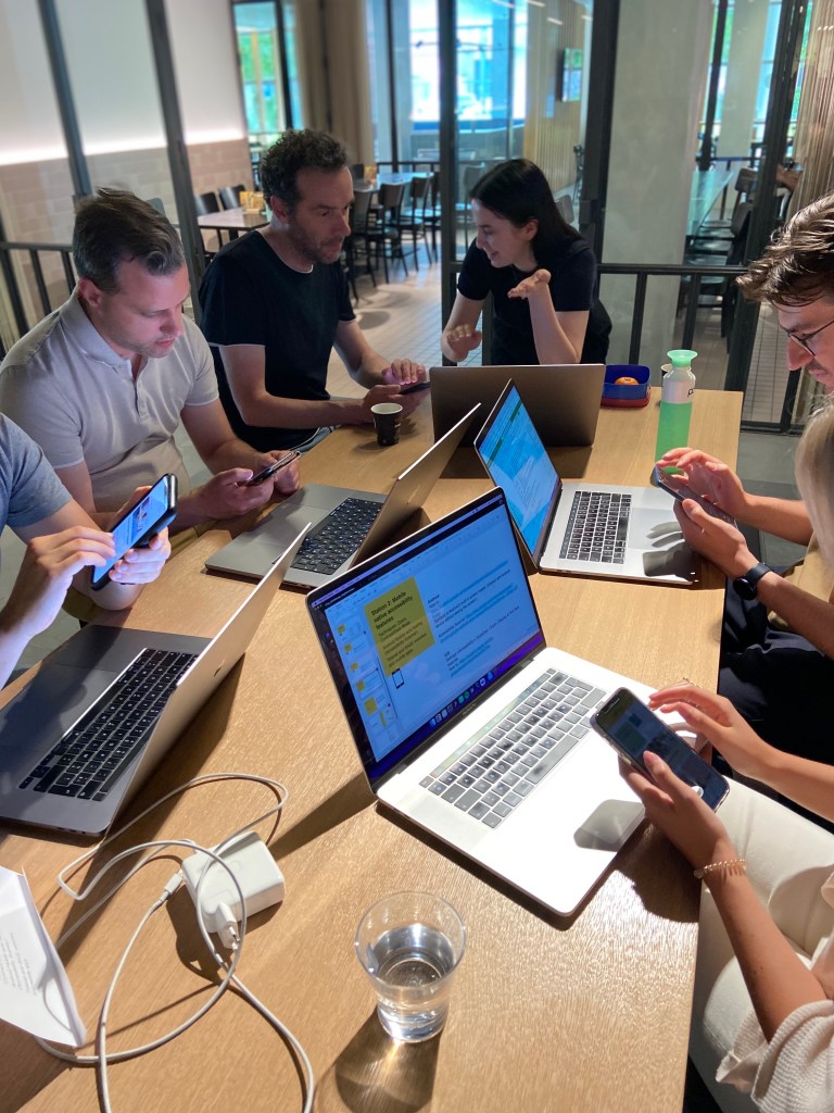 Group of seven people seating at a table facing their laptops and mobile phones testing.