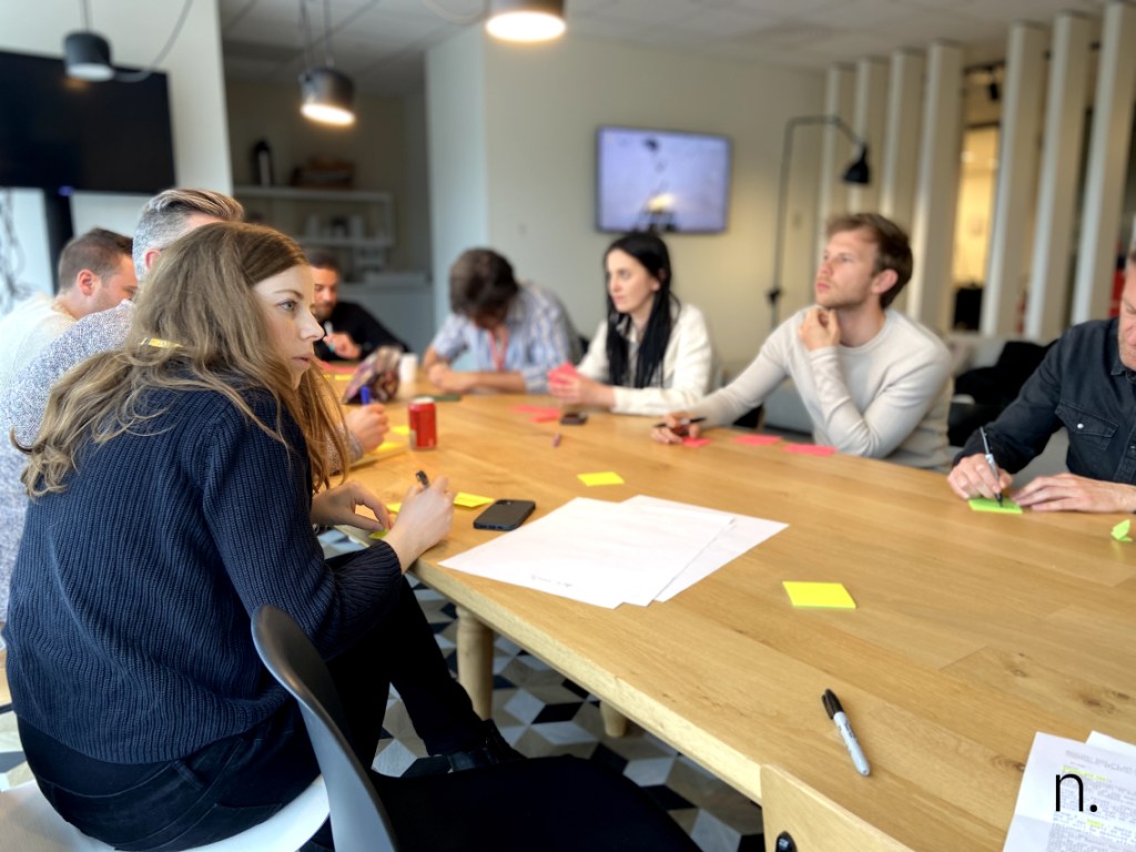 Group of people at a table with post its, markers and paper during a workshop. 8 visible people, focus on female in foreground.