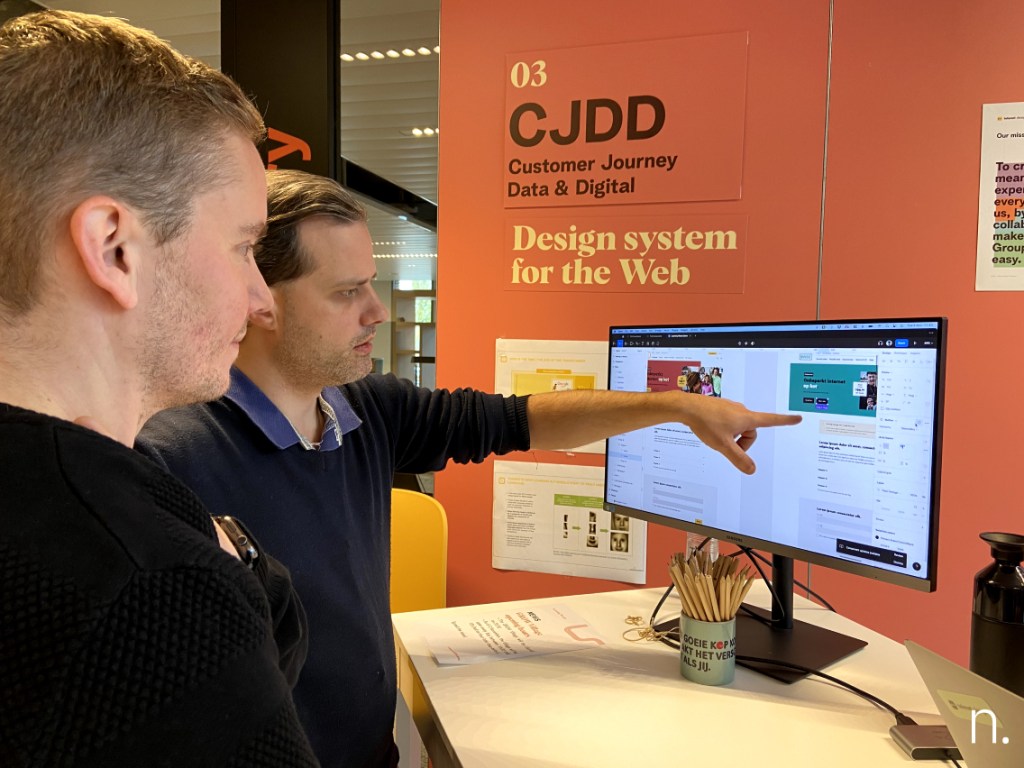 Two people, one pointing at a computer monitor. Reads in the background wall: CJDD Customer Journey Data & Digital Design System for the Web.