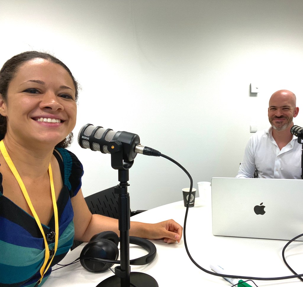 Nádia smiling recording a podcast with a standing microphone. Next to a men colleague using a Mac computer.