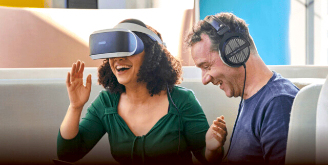 Nadia laughing and wearing VR glasses seating next to a men colleague wearing headphones.