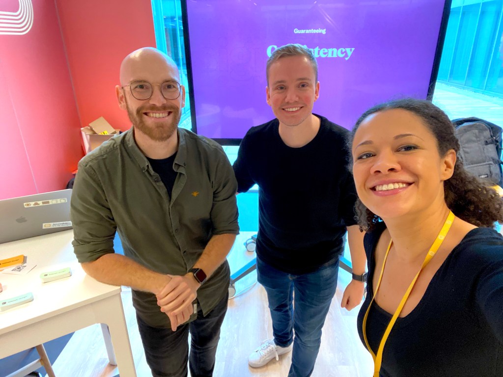 Selfie mode photo of Nádia with 2 other men colleagues smiling with a screen displaying “Consistency”.